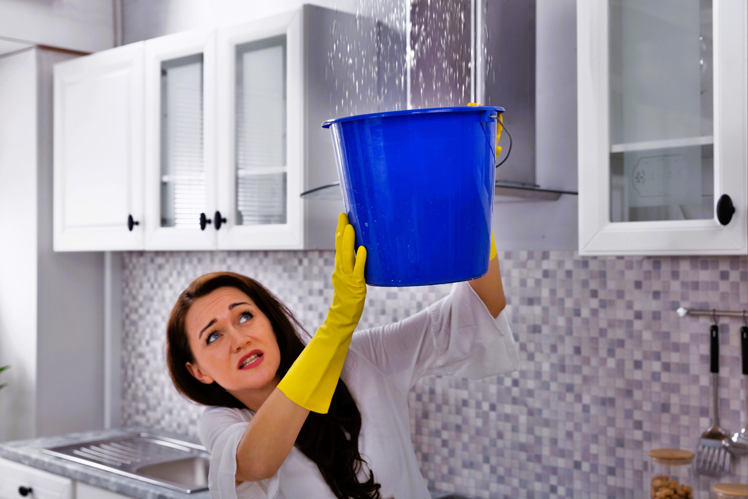 Unhappy Young Woman Collecting Water Leaking From Ceiling In Blue Bucket